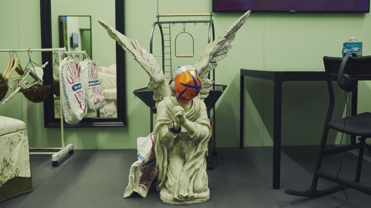 Installation view: a kneeling angel statue wearing a taped-up ball as a mask, in a pale green room with garment racks and a mirror